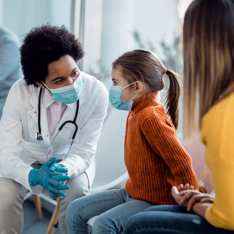 Doctor wearing a mask and gloves talks with a young girl patient in a clinic setting.