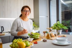 Smiling woman wearing glasses stands at a bright kitchen counter, chopping food with fresh fruit and herbs nearby in bowls and plants around her.
