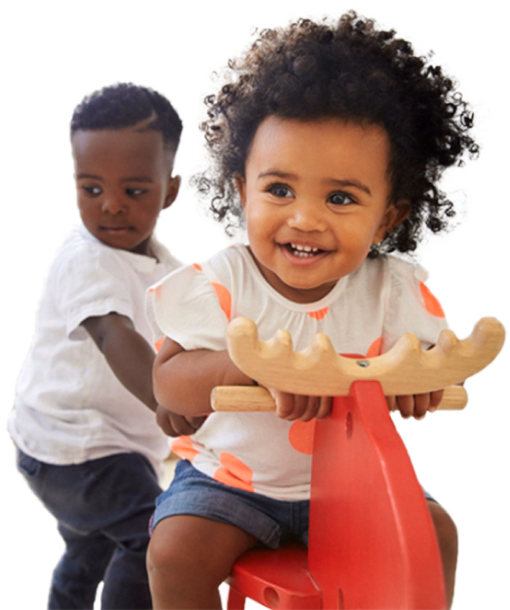 Two toddlers play on a red seesaw toy; the front-facing girl smiles while a boy stands behind her.