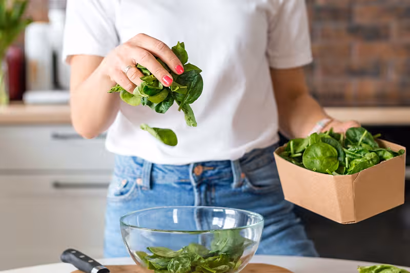 A person adds fresh spinach leaves to a glass bowl on a kitchen counter, pulling greens from a cardboard box nearby.