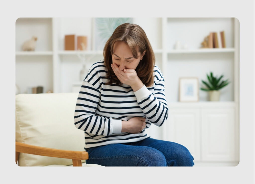 Woman sitting on a sofa, clutching her stomach and covering her mouth, appearing nauseated or in discomfort.",