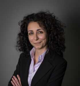 Portrait of a smiling woman with curly dark hair wearing a lavender shirt and black blazer, arms crossed.