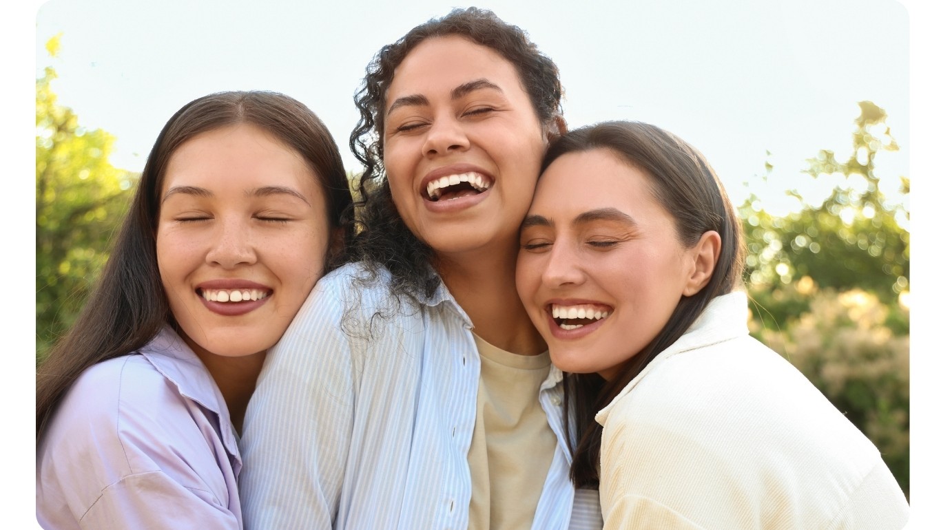 Three women smiling and hugging outdoors, showing friendship and joy in a sunny park setting
