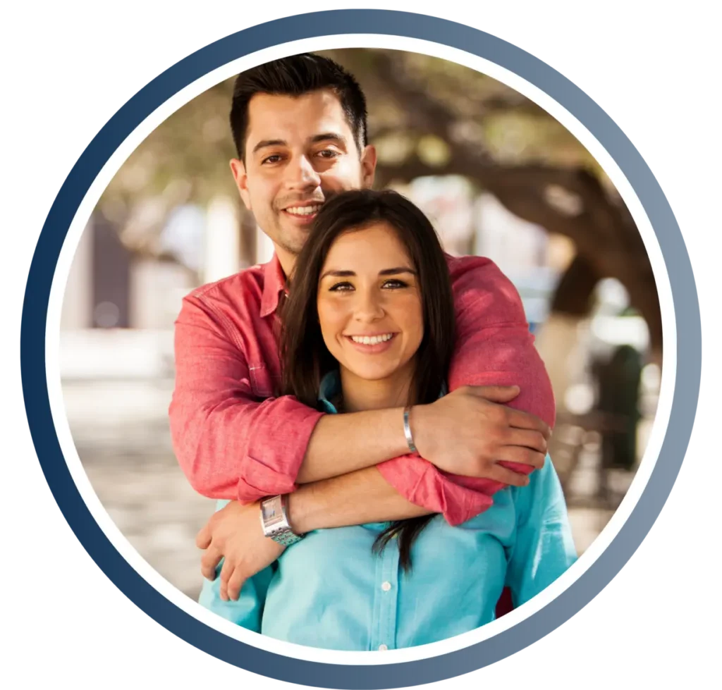 Smiling couple embracing outdoors, man behind woman with his arms around her shoulders in a park setting.