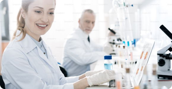 Smiling female scientist in a lab coat and gloves working at a computer in a bright laboratory.