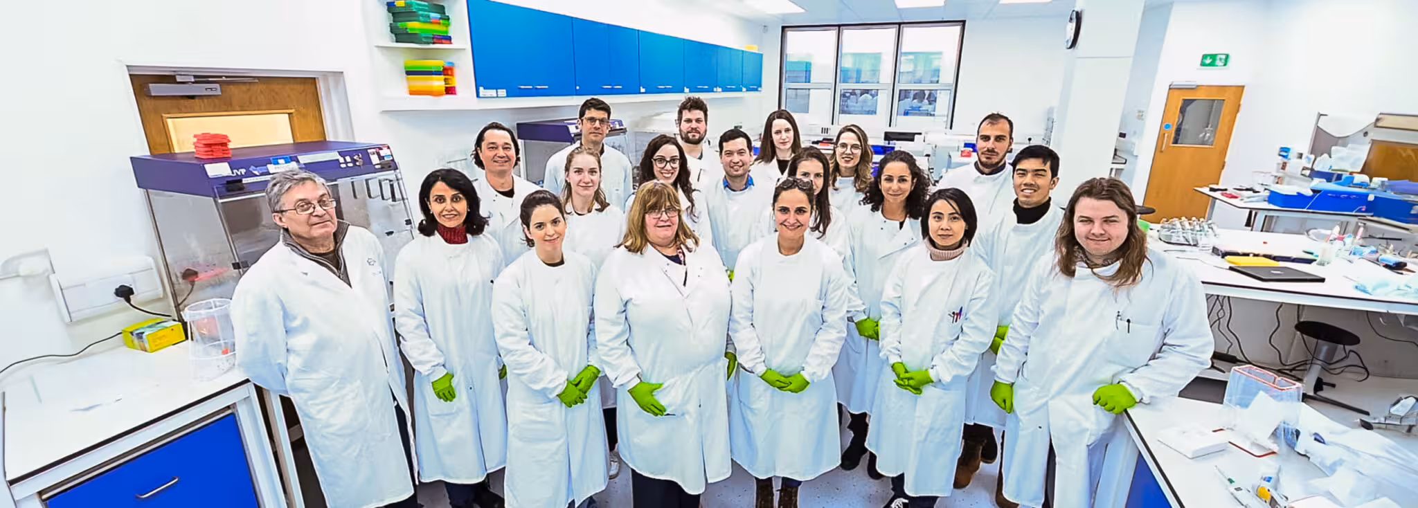 Group of scientists in white lab coats with green gloves posing in a bright laboratory with blue cabinets above them.