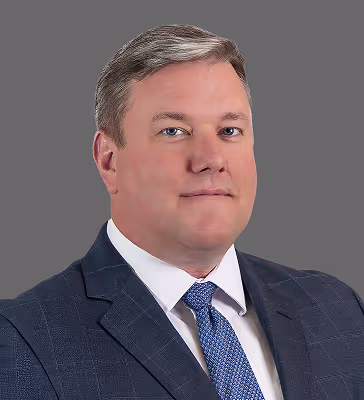 Portrait of a man in a dark blue suit and blue tie, against a gray background, looking at the camera.