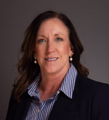Professional head-and-shoulders portrait of a smiling woman in a navy blazer and blue striped shirt against a gray backdrop. She's wearing pearl earrings.