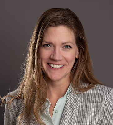 Professional headshot of a smiling woman with light brown hair, wearing a light blue blouse and gray blazer against a neutral gray backdrop.