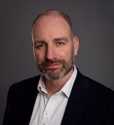 Professional headshot of a middle-aged man with a trimmed beard in a white shirt and black blazer against a gray studio background.