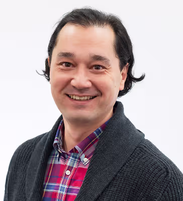 Smiling man facing the camera wearing a red plaid shirt and dark cardigan against a light background.