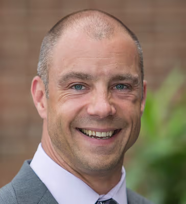 Smiling man in a gray suit and white shirt, outdoors, facing the camera.