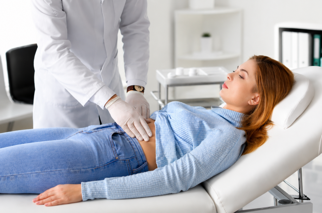 Woman lying on an examination chair while a clinician palpates her abdomen in a bright clinic.