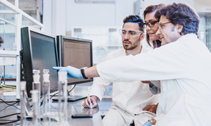 Researchers in a lab analyzing data on computer monitors, with one pointing at the screen while two colleagues observe