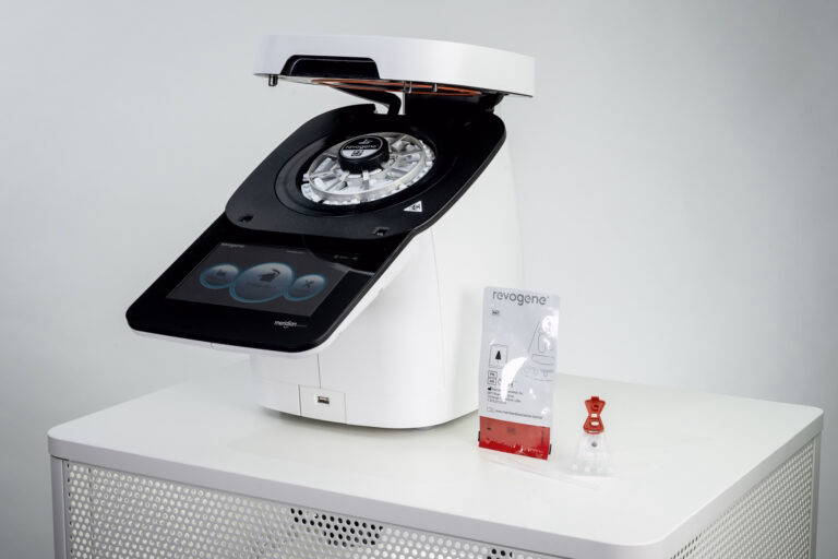 Laboratory molecular testing instrument on a white table with a touchscreen display and circular rotor, Revogene branding visible.