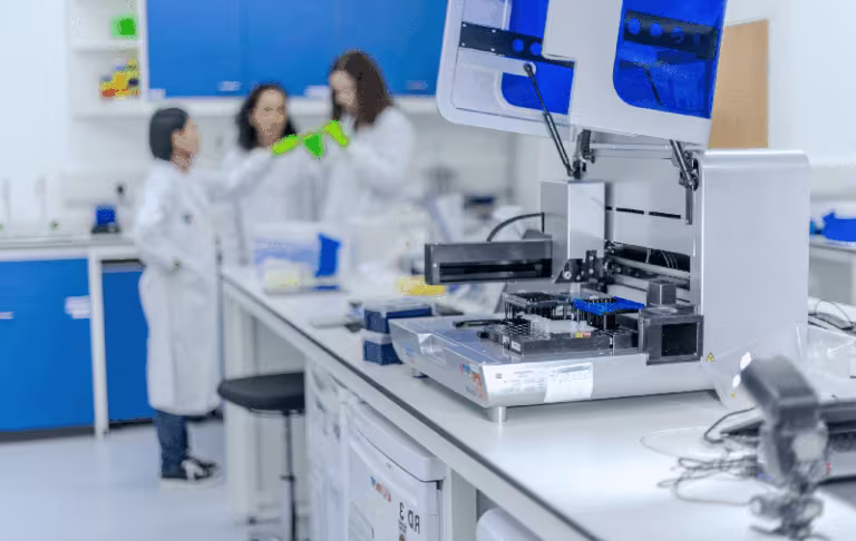 Three researchers in white lab coats stand at a clean laboratory bench around a large automated analyzer, with blue cabinets in the background; one pair of green gloves is visible as they discuss a sample